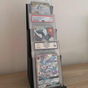 May include: Black tiered display stand holding graded Pok&eacute;mon trading cards. The cards are in protective cases, featuring characters like Charizard and Sylveon. The stand is on a wooden surface.