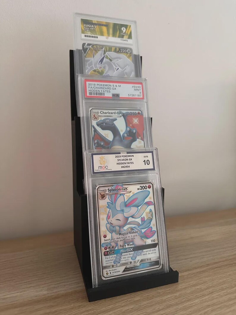 May include: Black tiered display stand holding graded Pok&eacute;mon trading cards. The cards are in protective cases, featuring characters like Charizard and Sylveon. The stand is on a wooden surface.