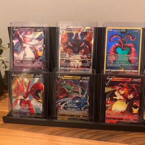 May include: A black display stand holding six Pok&eacute;mon trading cards in protective cases. The cards feature various characters with vibrant colors and unique designs. The stand is made of a dark material and sits on a wooden surface.