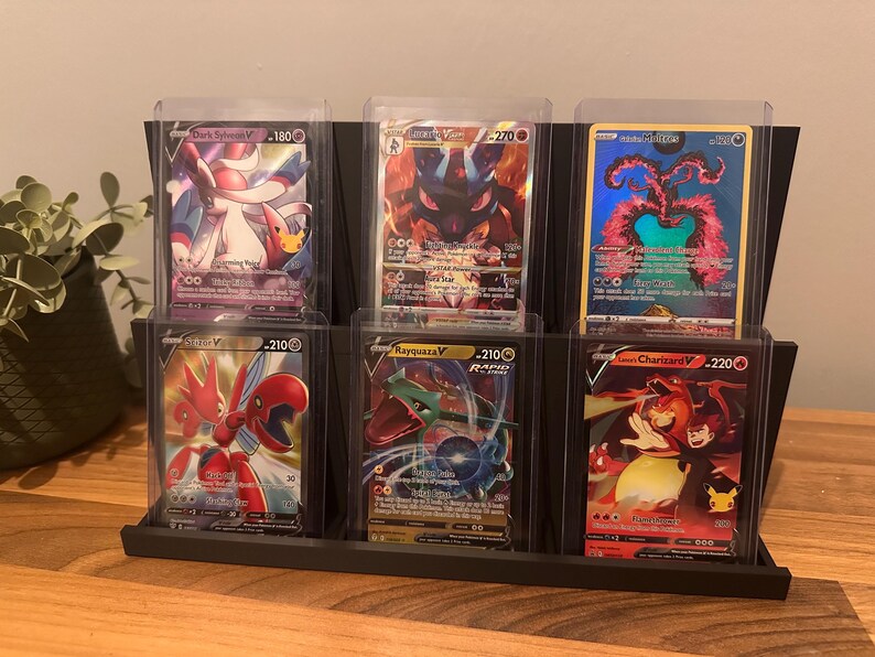 May include: A black display stand holding six Pok&eacute;mon trading cards in protective cases. The cards feature various characters with vibrant colors and unique designs. The stand is made of a dark material and sits on a wooden surface.
