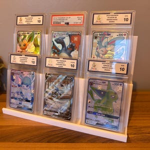May include: A white display stand holding six graded Pok&eacute;mon trading cards. The cards feature various Pok&eacute;mon characters, including Charizard, Sylveon, and Espeon. Each card is encased in a protective holder with a grade of 10.