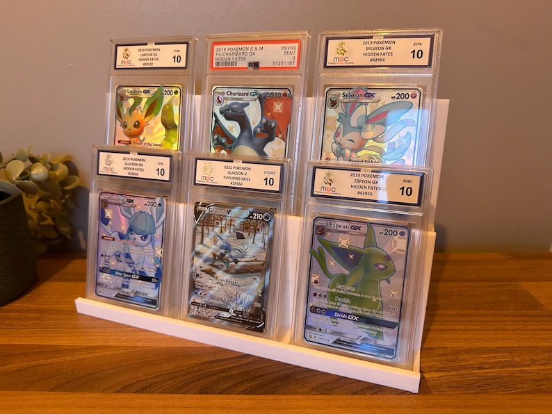 May include: A white display stand holding six graded Pok&eacute;mon trading cards. The cards feature various Pok&eacute;mon characters, including Charizard, Sylveon, and Espeon. Each card is encased in a protective holder with a grade of 10.