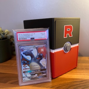 May include: A graded Charizard GX Pokémon card in a protective case, beside a red, white, and black storage box with a white "R" logo. The card is from the Hidden Fates set.