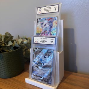 May include: A white display stand holding three graded Pok&eacute;mon trading cards. The cards feature Glaceon V and Glaceon VMAX from the Evolving Skies set. The cards are encased in protective holders with grading information visible.