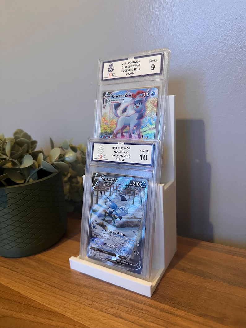May include: A white display stand holding three graded Pok&eacute;mon trading cards. The cards feature Glaceon V and Glaceon VMAX from the Evolving Skies set. The cards are encased in protective holders with grading information visible.