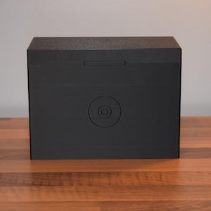 May include: A black rectangular box with a circular cutout on the front. The box has a smooth, matte finish.