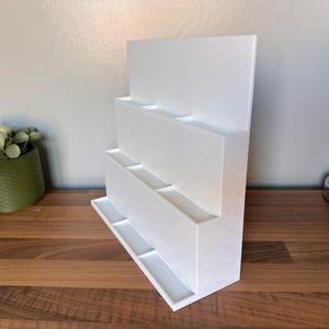 May include: A white, tiered display stand with multiple rectangular compartments. The stand has a flat base and a tall, vertical back. The design is modern and minimalist, suitable for displaying various items. The stand is made of a solid, opaque material.