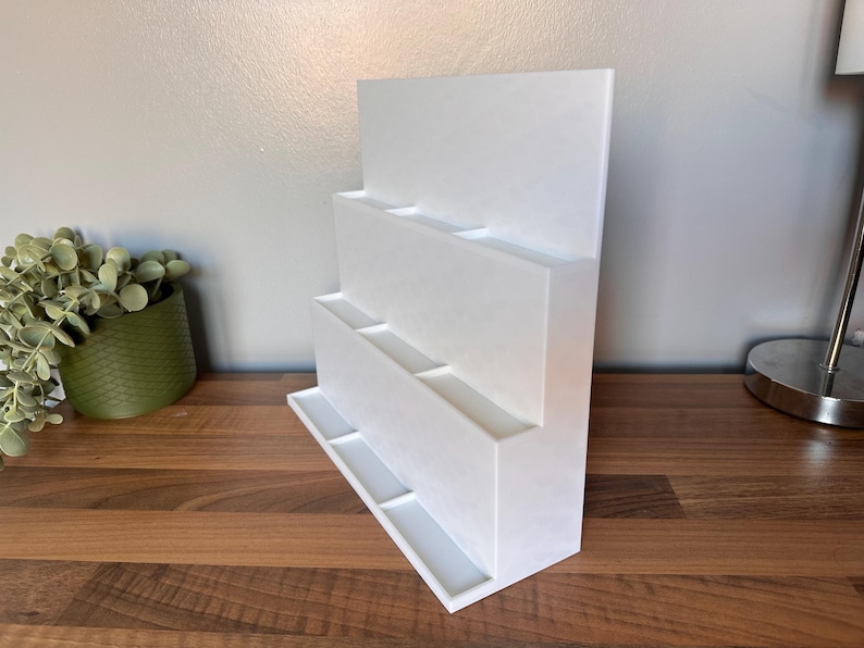 May include: A white, tiered display stand with multiple rectangular compartments. The stand has a flat base and a tall, vertical back. The design is modern and minimalist, suitable for displaying various items. The stand is made of a solid, opaque material.