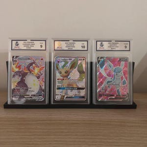 May include: Three graded Pok&eacute;mon trading cards displayed in protective cases. The cards feature Charizard VMAX, Leafeon GX, and Glaceon V. Each card is graded and encased in a clear plastic holder, with a black stand.