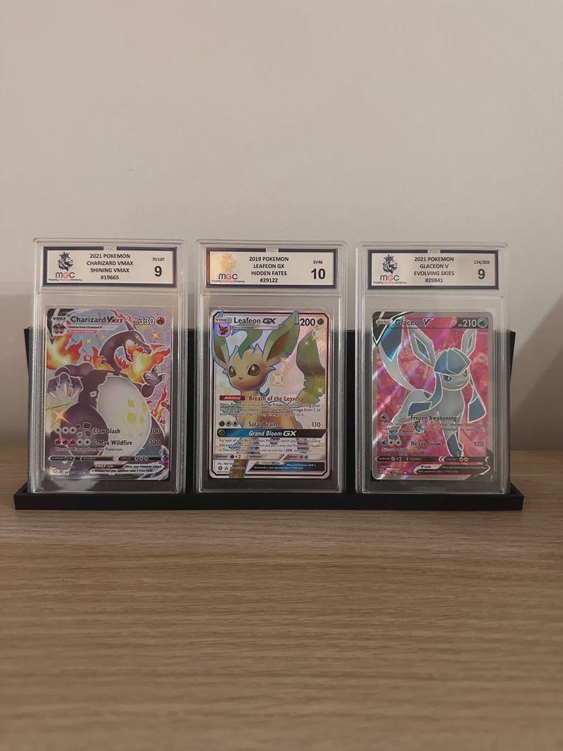 May include: Three graded Pok&eacute;mon trading cards displayed in protective cases. The cards feature Charizard VMAX, Leafeon GX, and Glaceon V. Each card is graded and encased in a clear plastic holder, with a black stand.