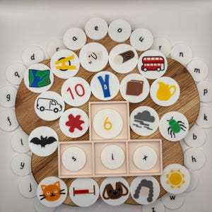 May include: A wooden board holds white circular tiles featuring lowercase letters, numbers, and colourful illustrations including a red bus, a yellow pitcher, a blue zipper, a bat, a cat, a dog, a sun, and a cloud, with a tray spelling out six.