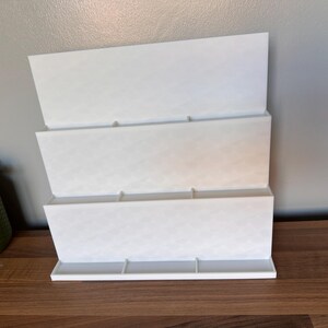 May include: A white, three-tiered organizer with a diamond pattern. The organizer is made of a solid material and has a rectangular base. It is designed to hold items and is placed on a wooden surface.