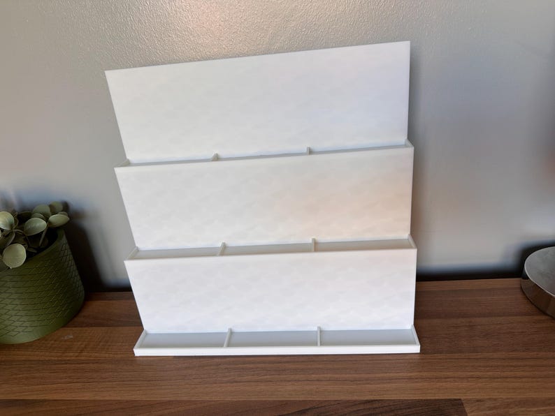 May include: A white, three-tiered organizer with a diamond pattern. The organizer is made of a solid material and has a rectangular base. It is designed to hold items and is placed on a wooden surface.