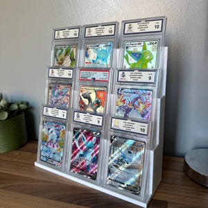 May include: A white tiered display stand holding graded Pok&eacute;mon trading cards. The cards are encased in clear plastic holders, showcasing various Pok&eacute;mon characters. The grades range from 9 to 10, with the text visible on the cards.
