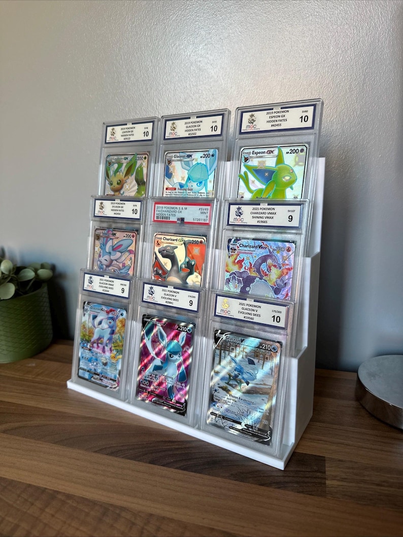 May include: A white tiered display stand holding graded Pok&eacute;mon trading cards. The cards are encased in clear plastic holders, showcasing various Pok&eacute;mon characters. The grades range from 9 to 10, with the text visible on the cards.