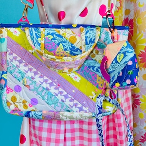 May include: A quilted tote bag featuring a diagonal patchwork design with vibrant floral, leaf, and small animal patterns in shades of blue, yellow, pink, and green. It includes a bright pink shoulder strap and a small matching quilted pouch.