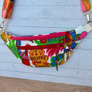 May include: A white fanny pack with a colorful floral and cartoon design, featuring the text "I Believe in Fairies." The bag has a red zipper and a pink interior. The strap is multi-colored with gold hardware.