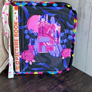 May include: A quilted picture book cover featuring a castle design in shades of pink, purple, and blue against a black background. The cover has a colorful geometric border and a white ribbon with pink stars. The spine reads "PICTURE BOOK".