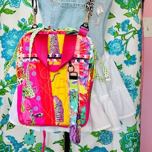 May include: A quilted backpack featuring a vibrant, multi-colored pattern with yellow, hot pink, and orange sections. The bag includes a tiger print, floral motifs, and fish graphics. It has bright pink top handles, a leopard print strap, and a wristlet.
