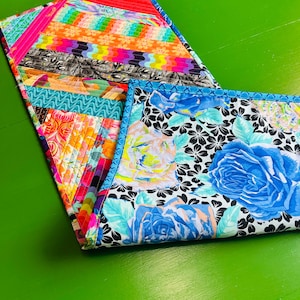 May include: A rectangular quilted table runner featuring a patchwork design of multicolored fabric strips in shades of orange, pink, yellow, and black, paired with a contrasting section displaying a bold blue rose floral pattern on a white background.