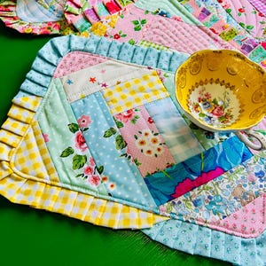 May include: A rectangular quilted placemat featuring a patchwork design of floral, gingham, and polka dot fabrics in pastel colors. The placemat is bordered by a pleated ruffle trim and is topped with a yellow teacup with a floral interior pattern.