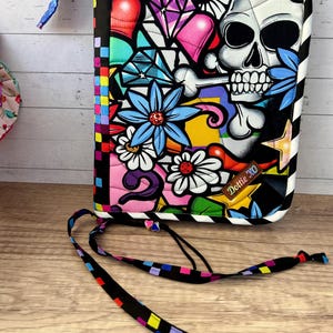 May include: A colorful, quilted case with a vibrant graphic design featuring a skull, flowers, hearts, and diamonds. The case has a black and white striped border and a matching strap with colorful square accents. The text "Dottie XO" is visible.