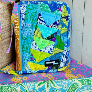 May include: A quilted, zippered case with a patchwork design in shades of blue, green, and yellow. The case features a variety of floral and geometric patterns. The text "I love you, I know" is on a small black label. A strap with the text "SPECK" is attached.