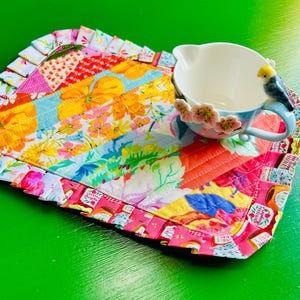 May include: A quilted fabric placemat featuring a vibrant floral pattern in shades of yellow, orange, and pink with a ruffled border, paired with a white ceramic mug adorned with small pink flowers and a small blue and yellow bird figurine on the rim.