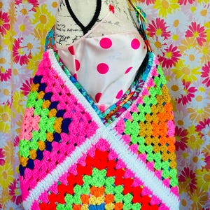 May include: A handmade crochet shoulder bag featuring a vibrant granny square pattern in neon pink, bright orange, lime green, yellow, and blue, accented by a white border. The bag has a wide strap and is constructed from colorful yarn squares.