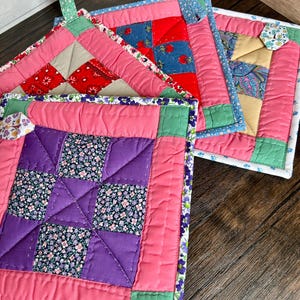 May include: A collection of colorful, quilted pot holders. The pot holders feature a patchwork design with various floral and geometric patterns in shades of pink, red, purple, blue, and green. Each pot holder has a loop for hanging.