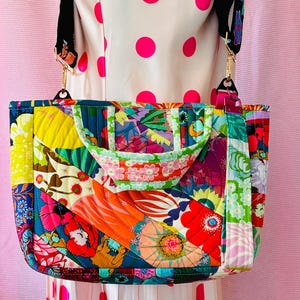 May include: A rectangular quilted tote bag featuring a vibrant patchwork design with various floral and abstract patterns in shades of red, orange, yellow, green, and blue. The bag includes two short handles and a detachable black patterned shoulder strap.