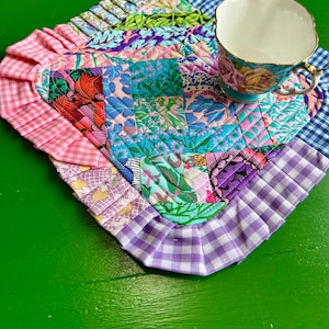 Puede incluir: Una alfombrilla de tela acolchada en forma de corazón con un mosaico de varios patrones florales y geométricos en tonos rosa, azul, morado y verde, rematada con un borde de volantes de cuadros vichy, coronada con una taza de porcelana floral.