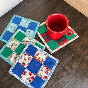 May include: A collection of handmade quilted coasters in various patterns and colors. One coaster features a red and green plaid design, while others showcase floral and patchwork patterns in blue, green, and white. A red mug sits atop one coaster.