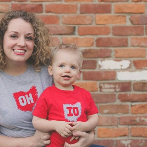 May include: A woman and a young boy are wearing shirts with the state outlines of Ohio and Indiana. The woman is wearing a grey shirt with the word "OH" in a red outline. The boy is wearing a red shirt with the word "IN" in a grey outline. They are standing in front of a brick wall.
