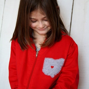 May include: A young girl wearing a red zip-up hoodie with a grey pocket featuring an outline of the state of Ohio and a small red heart.