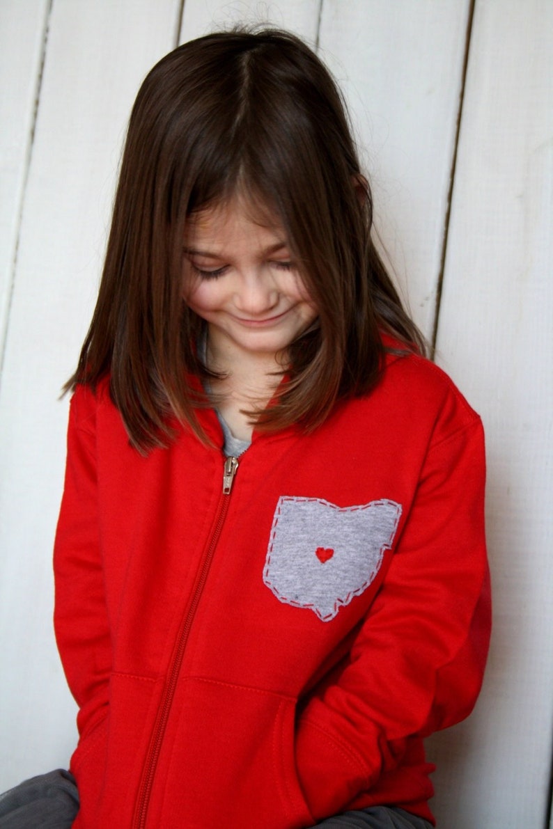 May include: A young girl wearing a red zip-up hoodie with a grey pocket featuring an outline of the state of Ohio and a small red heart.