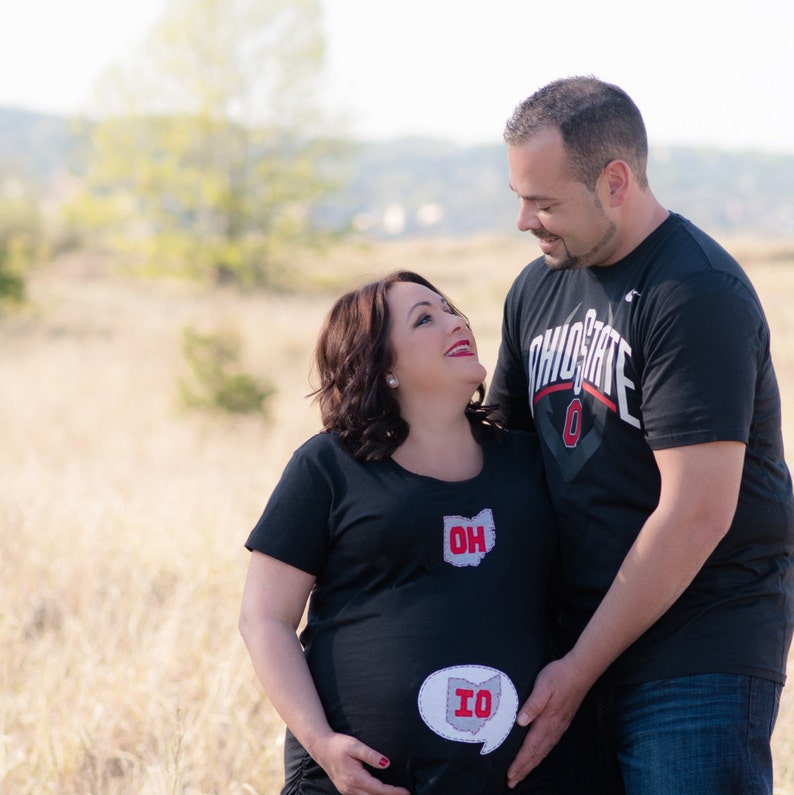 Pode incluir: Uma mulher gr&aacute;vida e um homem est&atilde;o vestindo camisas combinando do Ohio State. A mulher est&aacute; vestindo uma camisa preta com um contorno branco do estado de Ohio e as palavras "OH" e "IO" nela. O homem est&aacute; vestindo uma camisa preta com as palavras "Ohio State" e um grande "O" nela. Eles est&atilde;o em p&eacute; em um campo de grama alta.