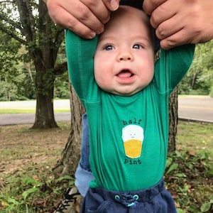 May include: A baby wearing a green long-sleeved shirt with a cartoon beer glass and the text "half pint". The baby is being held up by two hands.