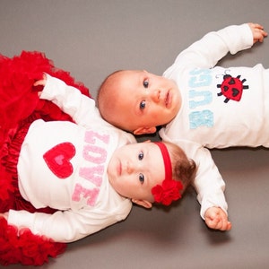 May include: Two babies wearing white onesies with embroidered designs. The baby on the left is wearing a red tutu and has a pink heart with the word "LOVE" on their onesie. The baby on the right has a ladybug with the word "BUGS" on their onesie.
