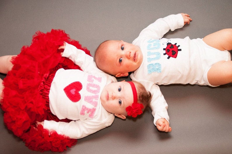 May include: Two babies wearing white onesies with embroidered designs. The baby on the left is wearing a red tutu and has a pink heart with the word "LOVE" on their onesie. The baby on the right has a ladybug with the word "BUGS" on their onesie.