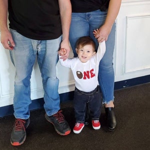 May include: A baby wearing a white onesie with a red and brown football design and the word "ONE" on it. The baby is being held by two adults, one wearing a black shirt and blue jeans and the other wearing a blue shirt and blue jeans.