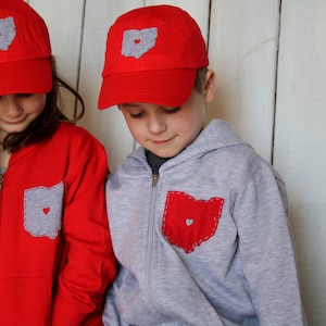 May include: Two children wearing red and grey sweatshirts with a grey and red Ohio state outline with a heart in the middle. Both children are wearing red baseball caps with a grey Ohio state outline with a heart in the middle.