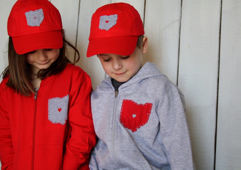 May include: Two children wearing red and grey sweatshirts with a grey and red Ohio state outline with a heart in the middle. Both children are wearing red baseball caps with a grey Ohio state outline with a heart in the middle.