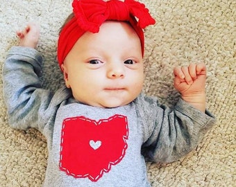 May include: A baby wearing a gray long-sleeved onesie with a red Ohio state outline and a small heart in the center. The baby is wearing a red bow headband.