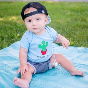 May include: A baby wearing a black baseball cap and a light blue onesie with a cactus graphic and the text "cuddle me". The baby is sitting on a blue blanket on a grassy surface.