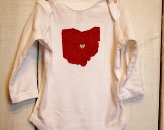May include: White long-sleeved baby onesie with a red glitter outline of the state of Ohio and a small white heart in the centre.