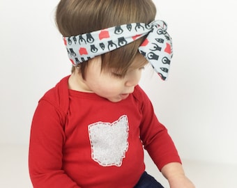 May include: A red long-sleeved bodysuit with a silver glitter Ohio state outline. The bodysuit is worn by a child wearing blue jeans with distressed knees and a gray headband with black and red handprint designs.