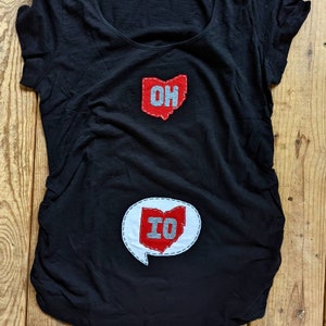 Pode incluir: Camiseta preta com patches de feltro vermelho em forma do estado de Ohio. O patch superior diz "OH" e o patch inferior diz "IO".