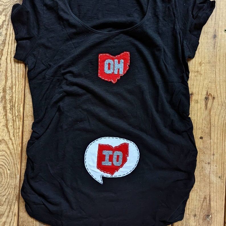 Pode incluir: Camiseta preta com patches de feltro vermelho em forma do estado de Ohio. O patch superior diz "OH" e o patch inferior diz "IO".