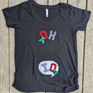 Pode incluir: Camiseta preta com um "OH" vermelho e cinza e um bal&atilde;o de fala branco com um "IQ" cinza em costura vermelha. As letras s&atilde;o decoradas com folhas verdes e bagas vermelhas.